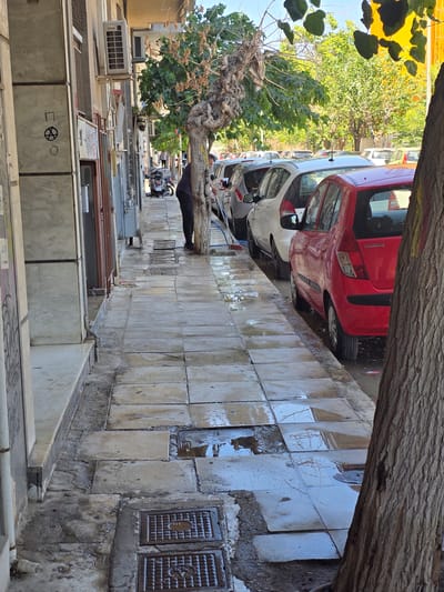 The image depicts a street scene in Athens, Greece, with a strong emphasis on parked cars and architectural elements. The foreground is dominated by a large tree trunk and its branches, casting shadows and partially obscuring the view. Beneath the tree, several cars are parked along the side of the street, including a prominent red Hyundai. The street appears to be paved with large, square tiles, many of which are wet and reflect the surrounding environment, suggesting recent rain or washing.
Several manhole covers are visible on the tiled sidewalk, indicating underground utilities. Adjacent to the street, there is a row of buildings with what appear to be air conditioning units mounted on the exterior walls. The architecture suggests a residential or commercial area with a mix of older and newer construction.
In the background, more trees and greenery can be seen, with sunlight filtering through the leaves. The overall lighting suggests it is daytime, possibly late morning or early afternoon, given the angle of the shadows. The presence of wet surfaces and the vibrant green of the foliage might indicate a time after a rain shower or a humid day.
There are no people clearly visible as active participants in a specific event, though a person can be seen in the background near a parked vehicle, possibly interacting with it. The scene conveys a sense of everyday urban life in Athens, characterized by its distinct architecture, street parking culture, and Mediterranean vegetation. The image captures a slice of the city's atmosphere, blending natural elements with urban infrastructure.