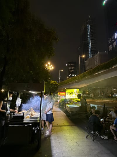 In a dimly lit urban setting, likely in Huangpu District, China, a street food stall is the focal point. The time appears to be night, indicated by the dark sky and artificial lighting. The stall is set up on a paved sidewalk next to a grassy median. A building with numerous illuminated windows is visible in the background, suggesting a commercial or residential area.
Several people are present. Two individuals are seated on chairs near the food stall, engrossed in their phones. Another person, wearing a white shirt and dark shorts, stands near the stall, seemingly waiting or observing. Further down the sidewalk, another person is seated, wearing glasses and a dark shirt. A vendor is actively preparing food at the stall, with smoke rising from the cooking area. Two individuals are also seen standing near the food stall.
The food stall itself is a prominent object, equipped with a display case for food items and a cooking area. The text visible on the stall and surrounding signage includes "老曾螺蛳粉", "牌仔燒烤", and "湿辣牛肉2". These indicate that the stall sells螺蛳粉 (Luosifen, a type of noodle soup) and烧烤 (shaokao, grilled skewers), specifically mentioning spicy beef skewers. The overall atmosphere suggests a typical evening street food scene, with people gathering to eat and socialize. The weather appears clear.