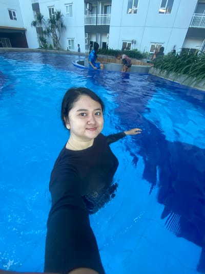 A woman in a black long-sleeved swimsuit is taking a selfie in a blue swimming pool, likely at an apartment complex in Sidoarjo, Indonesia. Several other people are standing at the edge of the pool. A multi-story building is visible in the background.