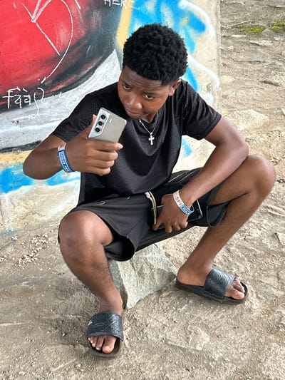 A young Black man is depicted in a rotated outdoor image, appearing to recline against a textured wall or sloped ground in Ihiagwa, Nigeria. He has short, curly black hair and is dressed in a black short-sleeved top, black shorts, and black slide sandals. A silver-colored cross necklace hangs from his neck.
He is holding a light-colored smartphone, featuring a distinct triple-lens camera setup on the back, in his right hand and looking down at its screen. On his left wrist, he wears two bands: a blue one with white text that reads "SFI IS REAL," and a white one with blue text partially visible, saying "VERY ACTIVE."
The background consists of a rough, light-colored concrete or stone wall, showing some signs of moss or discoloration. The ground where he rests is concrete, heavily adorned with colorful graffiti in shades of red, blue, black, and white, with some faint white script visible on the red section. The scene is brightly lit, indicating daytime and clear weather conditions. The man's expression is neutral as he engages with his phone, suggesting a moment of casual activity or relaxation in a public or semi-public outdoor space.