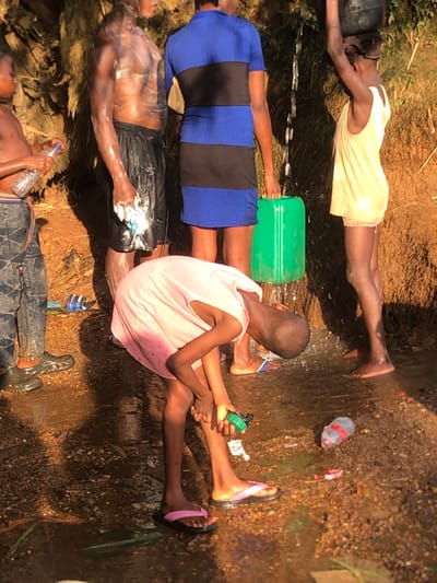 The scene depicts multiple individuals at an outdoor water source in Ukwu-Abwa, Nigeria. The ground is wet, muddy, reddish-brown earth with visible puddles and discarded plastic bottles and wrappers. A shirtless male, covered in white suds, stands in the center-left, wearing black shorts and holding a white cloth, suggesting a shower or wash. To his left, another young male, partially visible, wears patterned trousers and black Crocs while holding a clear plastic bottle. In the center-rear, a female in a blue and black striped dress is collecting water in a green plastic jerrycan from a flowing stream of water. To her right, a female in a light yellow top balances a dark container on her head. In the foreground, a young person in a pink t-shirt and pink flip-flops is bent over, washing or interacting with a small green object near their feet. The background shows an earthen bank with minimal vegetation, under direct sunlight.