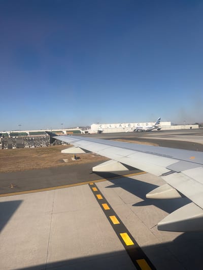 An airplane wing dominates the view, casting shadows on the tarmac with yellow markings. A building is labeled "C.L.A.G.D" with an airplane parked alongside. Various infrastructure and equipment are visible in the background, extending into the distance under a clear blue sky. The perspective suggests the photo was taken from inside an aircraft.