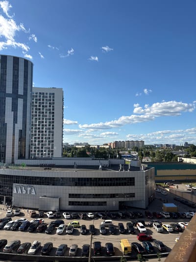 This is an aerial, landscape-oriented photograph taken from a high vantage point, looking down on an urban scene. The sky is a vibrant blue with scattered, elongated white clouds, suggesting a clear and sunny day. The time of day appears to be late morning or early afternoon, given the shadows cast by the buildings and cars.

The primary subjects are buildings and a parking lot filled with vehicles. Several modern buildings are visible, characterized by glass facades and geometric designs. One prominent building has a sign with the text "АЛЬТА" in large, white letters. Another building features a repeating pattern of windows. In the background, a densely populated residential area with numerous apartment buildings can be seen, interspersed with trees.

The parking lot in the foreground is occupied by a variety of cars, including sedans and SUVs, in different colors like black, white, silver, and red. The cars are parked in rows, indicating a busy commercial or residential area. There are a few people visible in the parking lot, some walking near the buildings. Some wooden crates are also present in the parking lot. The overall scene depicts a bustling urban environment in Mytishchi, Russia.