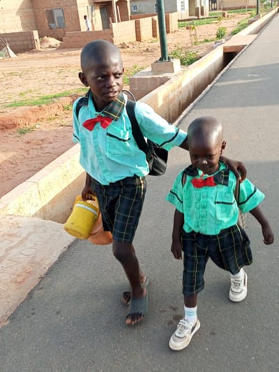 Two young boys, dressed in matching light blue short-sleeved shirts with red bow ties and plaid shorts, walk along a paved road in Tempure, Nigeria. The older boy on the left, carrying a yellow container in his right hand and a black backpack on his back, has his left arm around the shoulders of the younger boy on the right, who also wears a black backpack. The younger boy looks down with a sad expression. The environment includes a dry, dusty area with some vegetation to the left of the road and a concrete drainage ditch to the right. In the background, there are low-lying brick buildings. The lighting suggests it is daytime, possibly late afternoon, with long shadows cast on the ground.