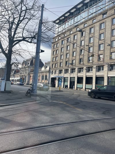 This image presents a daytime urban street scene in Geneva, Switzerland, likely captured from inside a vehicle due to visible reflections on the lower portion of the frame. The setting suggests winter or early spring, as a large deciduous tree in the foreground has bare branches.
In the immediate foreground, two sets of tram tracks run across an asphalt road, curving towards the left. A tall, grey utility pole stands prominently on the left, supporting a complex network of overhead power lines used by the city's trams. A bicycle is leaning against the base of this pole.
Along the right side of the street, a black SUV is parked. Further down the road, several people are walking on the sidewalk, and a blue and white tram is partially visible in the distance.
The street is flanked by diverse architecture. On the far left, a modern, multi-story building with a reflective glass facade stands tall. Adjacent to it, and dominating the right side of the frame, is a multi-story building with a more traditional, stone-like facade featuring numerous rectangular windows. Below this building, a blue sign with the text "AXA" is discernible. In the background, hints of an older, ornate building with classical architectural details can be seen.
The weather appears clear and sunny, indicated by the bright sky and distinct shadows. A small yellow sign displaying the number "15" is attached to one of the overhead wires, possibly denoting a tram route. The scene depicts a typical busy city environment with both contemporary and historical elements.