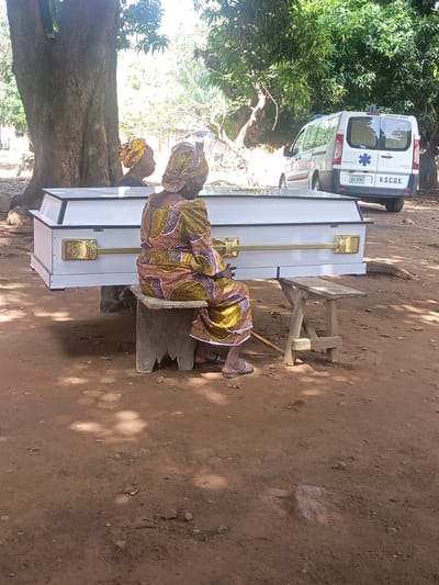 The scene is outdoors in a rural area, likely in or near Ninzo South, Nigeria, judging by the vehicle license plate. The ground is reddish-brown dirt. A white coffin with gold-colored handles and hinges rests on two uneven benches. A woman in a patterned dress and head wrap is sitting on one of the benches, facing the coffin. A second woman with a head wrap is partially visible behind the coffin, near the left side of the frame. An ambulance is parked in the background, its rear facing the camera. Trees are visible in the background, providing some shade.
