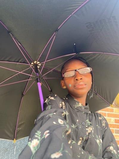 A low-angle, close-up photograph captures a young individual with dark skin, wearing glasses with reddish-purple frames, and braided hair. A small, light blue star-shaped sticker is visible on their right cheek, and they wear gold-colored infinity symbol earrings. They are dressed in a black, long-sleeved, collared shirt with a floral pattern featuring light-colored blossoms and green foliage. The individual holds a black umbrella with purple ribs and a purple handle open above their head. Faint, inverted white text is visible on the inner surface of the umbrella canopy. A light brown brick wall forms part of the background, while a gray, diamond-plate textured surface is visible below the umbrella. The specific location within Iranla, Nigeria, cannot be determined from the visual content.