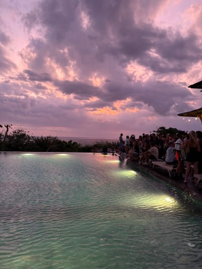 The image depicts an outdoor evening scene at an infinity pool facility in Uluwatu, Indonesia, during twilight hours. The sky above is characterized by an extensive cloud cover with pronounced purple and pink hues, indicative of a sunset, interspersed with darker grey cloud formations. The horizon line separates the sky from a calm, dark-blue ocean visible in the mid-ground.
In the foreground, a large, light-blue infinity pool occupies the lower left portion of the frame, showing ripples on its surface. Four submerged lights within the pool emit distinct yellow-green glows, illuminating the water. To the immediate right of the pool, a wooden deck extends, accommodating a gathering of approximately 30-40 individuals. These individuals are predominantly standing in various groups, engaged in social interactions, while some are seated on wooden lounge chairs or benches. A few individuals are visible further back, closer to the ocean's edge, also gathered socially. One person stands near the far edge of the infinity pool, facing the ocean. On the far right, a portion of a building or canopy roof, constructed from a natural material and adorned with string lights, is visible. Lush green vegetation borders the scene on the left, obscuring part of the ocean view.