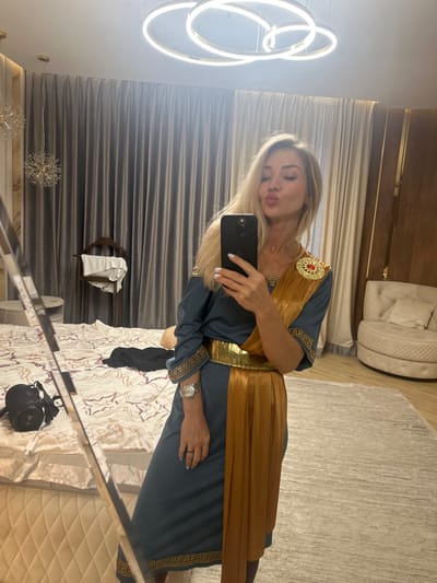 A woman is taking a selfie in a bedroom. She is wearing a blue and gold dress with a large gold belt and a brooch on her shoulder. She is puckering her lips for the photo. The room is decorated in a modern style with large curtains, a tufted armchair, and a large abstract light fixture on the ceiling. There is a bed with a patterned duvet and some clothes on it. A floor lamp with a crystal lampshade is visible in the background. The woman is standing on a light-colored rug. The image does not contain any recognizable landmarks or features that would specifically place it in Armavir, Russia; it is a generic bedroom interior.
