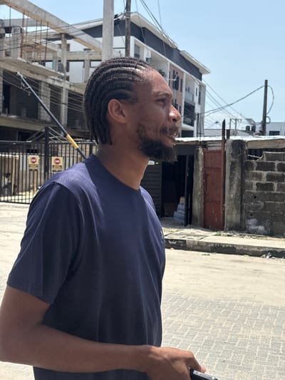 A man with cornrows and a beard stands on a paved surface in Igboefon, Nigeria, looking upwards. He wears a navy blue t-shirt and holds an object in his right hand. Buildings are visible in the background, including a multi-story structure under construction and an older, weathered building with a rusted metal door. Overhead power lines crisscross the sky. A metal fence with warning signs is also visible.