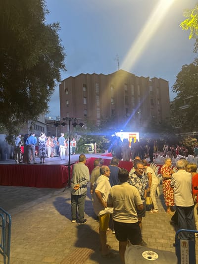 The image shows a group of people gathered outdoors, likely for an event or performance. Several individuals are seated in chairs, while others stand. The scene appears to be a street or plaza, with buildings visible in the background and trees lining the area. A stage with bright lights is set up, suggesting a performance is underway or about to begin. The lighting and attire of the people suggest it is evening or late afternoon. The overall atmosphere seems casual and social. There is a blue cart with text on it, partially visible, which may indicate a specific venue or organization. The location is identified as Alicante/Alacant, Spain.