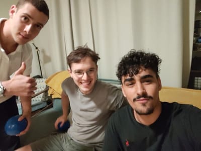 The image depicts three young adult males indoors in West Jerusalem, Israel. The male on the left, with short dark hair, wears a white collared shirt and a silver wristwatch on his left wrist. He presents a "thumbs up" gesture with his left hand and holds a blue spherical object in his right hand. The central male, with short brown hair and circular eyeglasses, is seated and smiles broadly towards the camera, wearing a grey t-shirt and olive-green shorts. A second blue spherical object is partially visible near his left leg. The male positioned closest to the camera on the right has dark curly hair, a mustache, and a beard, wearing a black t-shirt with a small red embroidered logo, and looks directly at the camera with a neutral expression. The background consists of light-colored curtains, a mustard-yellow fabric object draped over seating furniture, and a dark, slender object, consistent with a lamp stand, partially visible on the left.