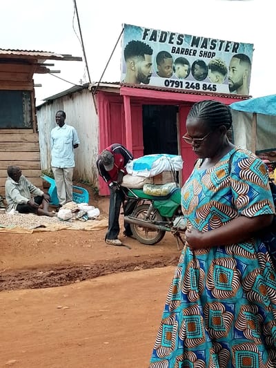 The image shows a street scene in Lwandeti, Kenya (latitude: 0.563, longitude: 34.806), with a woman in the foreground, a barber shop sign in the background, and several men in between. The woman is wearing a blue and gold patterned dress, and she is standing on a dirt road. Behind her, to the left, a man in a blue shirt is standing. He appears to be looking at something off-screen, out of frame. Behind him, to the left, is a man seated on the ground, holding a white object in his lap. To the right of the woman, a man wearing a red shirt and black pants is standing near a motorcycle. The motorcycle has a blue tarpaulin, and is being loaded with other bags. There are bags on the ground near the motorcycle, and a few other items, but it is not clear what they are. The man in the red shirt appears to be putting something into a bag, and the motorcycle appears to be parked, as the rider has stepped off of it. There is a sign above the motorcycle that says "Fades Master Barber Shop" with a phone number underneath. The sign has several pictures of men with different hairstyles. It is unclear what the man in the red shirt is doing. The woman is facing towards the camera, while the three men behind her are all facing to the left. There is a wooden structure behind the men, which may be part of the barber shop. The street is a dirt road, with a muddy patch near the motorcycle. It is a sunny day.