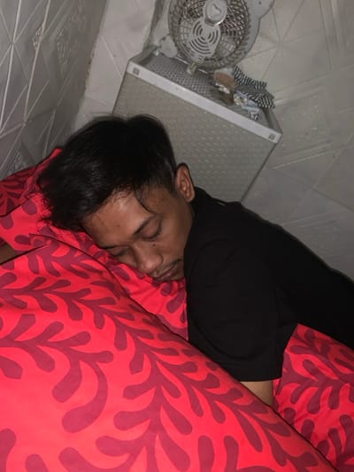 A person sleeps on a red pillow with a coral design. They are wearing a black top. Beside them is a white woven plastic storage unit. A small white fan sits on top of the unit, with some items placed behind it. The floor is tiled in a white geometric pattern. The picture has a slightly rotated perspective.