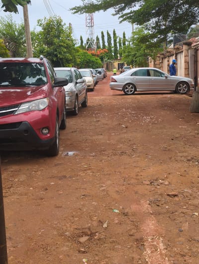 The image displays a line of cars parked on the side of a reddish-brown dirt road in what appears to be a residential or semi-rural area, consistent with Unguwar Rimi, Nigeria.
The closest vehicle is a red SUV or crossover, with visible raindrops on its surface, indicating recent rain. Behind it, a silver sedan, identifiable as a Mercedes-Benz C-Class (W203 generation), is parked. Several other vehicles, mostly light-colored, are visible further down the line.
The road itself is unpaved, made of reddish-brown earth and mud, appearing wet and uneven in several spots, with scattered small stones and debris. To the left of the parked cars, a dense line of green trees and shrubs forms a natural barrier, with a light-colored wall or fence visible behind them. A dark water tank is mounted on a structure further back. In the background, a prominent red and white lattice communications tower stands tall against the sky.
One person wearing a blue shirt and cap is partially visible standing near the silver Mercedes-Benz in the distance. Another individual is faintly discernible near the front of the red car, somewhat obscured by the trees.
The scene takes place during the daytime, likely under overcast conditions or following rainfall, given the wet appearance of the cars and ground. There is no discernible text present in the image.