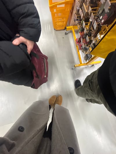 A high-angle shot showcases a white floor where three people stand. One wears olive green cargo pants and black shoes, while another dons a light gray coat and mustard yellow loafers. The third person wears a black jacket and stands next to a maroon backpack. A yellow cart filled with various items is visible in the background.