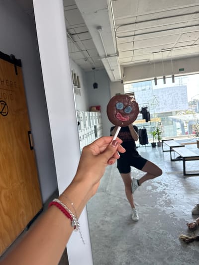 A person is holding a chocolate lollipop with blue and red candy decorations in front of a mirror, seemingly taking a selfie. The reflection in the mirror shows a person lying on the floor in black shorts and white sneakers. The location appears to be inside a building with large windows, potentially a studio or commercial space. In the background, through the windows, there are blurred urban elements suggestive of an outdoor environment. Based on contextual clues like the presence of a sign that says "Studio" on the furniture, the image may be a studio space in Puebla, Mexico. The lighting is predominantly bright, natural light coming through the windows.