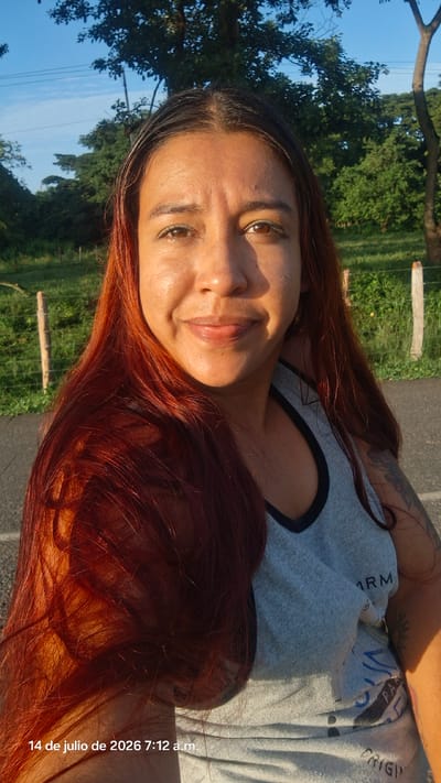 A medium shot shows a woman from the chest up, facing the camera. She has long, reddish-brown hair that cascades over her shoulders and chest. Her skin is tanned, and she is smiling with her lips slightly parted. She is wearing a light gray tank top with black trim around the neckline and armholes. The tank top has some text and designs on it, but they are partially obscured. On her left arm, a tattoo is visible.
The background is a natural outdoor setting. To the left and behind her, there is a grassy area with wooden fence posts. Further back, there are trees and foliage under a clear blue sky with some wispy clouds. To the right, there is a paved surface, likely a road, and more greenery. The lighting suggests it is daytime, possibly morning or late afternoon, given the golden hue on her skin. The date and time stamp in the bottom left corner reads "14 de julio de 2026 7:12 a.m." indicating the image was taken on July 14, 2026, at 7:12 AM. The location is identified as Vallecito, Venezuela.