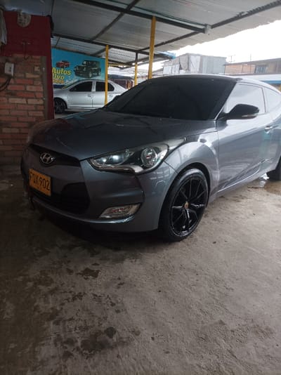 The image displays a gray Hyundai Veloster hatchback parked indoors, likely within a covered car wash bay or a garage. The car is positioned slightly angled towards the viewer, highlighting its front and right side. It features a clean, glossy finish, dark tinted windows, and distinctive black multi-spoke alloy wheels. The headlights are illuminated, and small fog lights are visible in the lower bumper. A yellow license plate, "FOX 902", is mounted on the front bumper.
The floor beneath the car is concrete, showing patches of dampness and general wear, suggesting a functional area for vehicle cleaning or service. To the left, a reddish brick wall is visible, with a white electrical box or switch attached to it. In the background, behind the main car, a bright blue banner or sign is displayed, advertising "Autos Dúo" in white text, accompanied by illustrations of a red car and a classic dark green Jeep-like vehicle. Further back, a silver sedan is partially visible, parked behind the banner.
The area is covered by a corrugated metal roof supported by yellow metal beams. The lighting is diffuse and even, indicating an overcast day or ambient light filtering into the covered structure. The scene is devoid of people, focusing solely on the vehicles and their immediate environment.