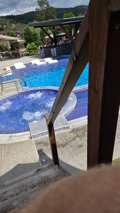 The image shows a partially visible person's leg in the foreground, obscuring a view of an outdoor swimming pool and its surroundings. The pool is tiled in blue and white, with a section of the pool appearing to be a jacuzzi with bubbling water. There are lounge chairs by the pool, and a bar area with bottles of alcohol is visible in the background. Beyond the pool area, a lush green forested hillside slopes upwards, with a few buildings nestled amongst the trees. The sky is visible and appears cloudy, suggesting it might be a partly cloudy day. The scene is set in Bania, Bulgaria, and the lighting suggests it is daytime, likely during the warmer months. The overall impression is a relaxing resort or leisure setting. Some text is visible on a purple refrigerator, which appears to say "Milka".