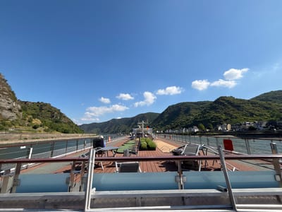 This image depicts the deck of a river cruise ship, likely on the Rhine River, as it passes through the scenic landscape near Kestert, Germany. The sky is a clear, vibrant blue with a few scattered white clouds. Lush, green, tree-covered hills rise on either side of the river, with a small German flag visible on a mast towards the rear of the ship. The deck itself is made of reddish-brown wood, with several lounge chairs and tables set up. Some potted plants are also visible. In the foreground, a metal railing and part of the ship's structure are prominent. The river is calm and dark, with a stone embankment visible on the left side of the image, and a town with white houses nestled on the hillside on the right.