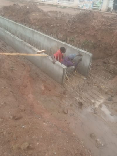 This image depicts an outdoor construction scene in Kakuri Gwari, Nigeria. Two individuals are actively engaged in building a concrete structure, possibly a drainage system or foundation. One person, wearing a red shirt, is standing and appears to be supervising or assisting. The other, dressed in a purple shirt and headscarf, is kneeling and working on the concrete wall, possibly cementing or reinforcing it. The surrounding environment is muddy and disturbed earth, indicating recent excavation or construction activity. Reinforcement bars are visible protruding from the concrete structure and extending into the ground. A wooden plank rests on the edge of the concrete. The overall scene suggests ongoing civil engineering work in a developing area.