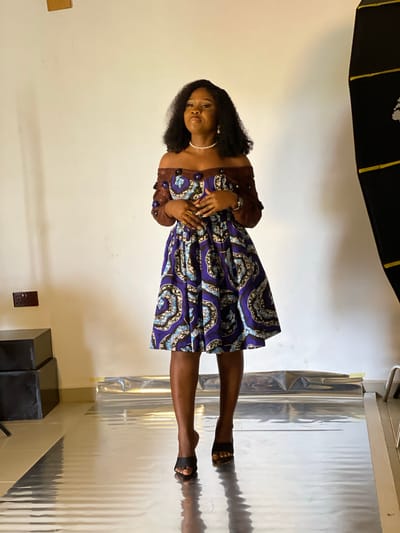 The image shows a young Black woman posing in a studio setting. She is wearing a brown jacket over a blue and white patterned dress and black sandals. Her hands are clasped in front of her. She has dark, curly hair and is wearing a pearl necklace. The background is a plain white wall, with some metallic sheeting on the right side of the frame. There is a black object, possibly a speaker, in the lower right corner. The lighting suggests an indoor environment, possibly a photoshoot. The woman has a neutral expression.