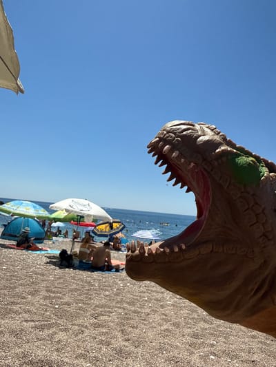 A plastic dinosaur toy, with its mouth open and teeth bared, is positioned in the foreground of a beach scene in Kamenovo, Montenegro. The toy, brown with some green accents, dominates the right side of the frame. Behind it, a pebble beach is visible, populated by beachgoers sitting under various umbrellas and tents. The sea is calm and blue, with a clear sky overhead. People are seen relaxing, some in swimwear, and a few are partially obscured by the beach umbrellas. The overall atmosphere suggests a sunny day at the beach.