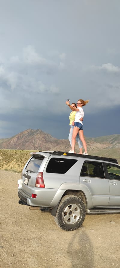 Two women are standing on the roof of a silver SUV in a rural, mountainous area. The woman on the left is wearing a yellow top and blue jeans, and the woman on the right is wearing a white t-shirt and denim shorts. The woman on the right is holding a smartphone and taking a selfie. The SUV has a roof rack and off-road tires. The background consists of barren, rolling hills and mountains, with a dramatic, cloudy sky. The location is identified as Lanjazat, Armenia.