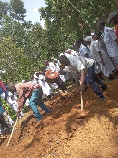 The image shows a burial taking place in a clearing in a wooded area in Malava, Kenya. The coordinates provided (latitude: 0.525, longitude: 34.814) are in the region of Malava, Kenya.  There is a group of people gathered around a freshly dug grave. One man is using a shovel to fill in the grave, while others stand around or observe the scene. The scene is in a wooded area with leafy green trees and shrubs.  There is a large wooden drum in the background, and several individuals are wearing white clothing, possibly religious or ceremonial attire. The atmosphere appears somber and respectful.