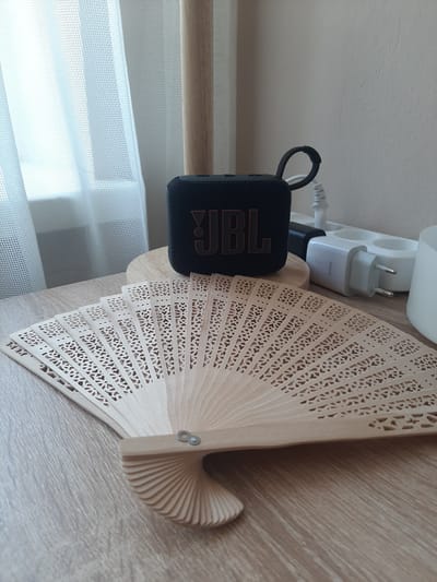 A close-up, high-angle, outdoor shot shows a wooden hand fan, fanned out on a wooden table, with a black JBL speaker and a white electrical outlet with plugs behind it. The fan is made of light-colored wood and features an intricate, laser-cut design. The JBL speaker is black with orange JBL lettering. Behind the speaker, a white power strip is visible, plugged into which are two white wall adapters and a black plug. The background consists of a sheer white curtain, partially obscuring a window, and a light-colored wall. The scene appears to be in an indoor setting, likely a room.