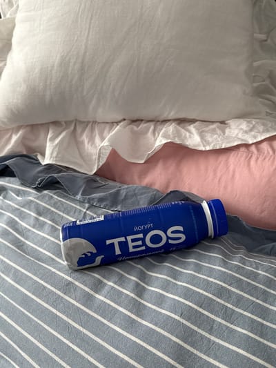 A blue bottle of TEOS yogurt lies on a bed with blue and white striped sheets. The bed has pillows covered in white and pink fabric, and a ruffled white bed skirt. The bottle is oriented diagonally across the frame, with its cap facing the top right. The label on the bottle is predominantly blue and white, featuring the brand name "TEOS" in large white letters and some Russian text. The scene appears to be indoors, likely a bedroom. The location of Prikubanskii, Russia is not visually discernible from the image.