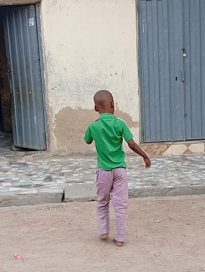 The image depicts an outdoor urban scene in Gwagwarwa, Nigeria, featuring a single, young, dark-skinned individual viewed from behind. The individual, appearing pre-adolescent with a shaved head, wears a bright green short-sleeved polo shirt and light purple/pink long pants, and is barefoot. They are in motion, walking away from the camera, with arms slightly bent and their head turned slightly to the left.
The foreground consists of unpaved sandy earth, on which a small, red, rectangular discarded object is visible in the lower-left corner. Immediately behind this, the ground transitions to a sidewalk or pathway paved with irregularly shaped light-colored tiles set in mortar.
In the background stands a light-colored building wall, showing significant wear, including peeling paint and darker moisture-related discoloration. Faint light-colored markings and text, resembling graffiti or notes, are present on the upper section of the wall. Two dark gray corrugated metal doors are integrated into this wall: the door on the left is partially open, revealing a dark interior, and has a small sticker on its left edge; the door on the right is closed and secured with a visible padlock. The scene is illuminated by natural daylight.