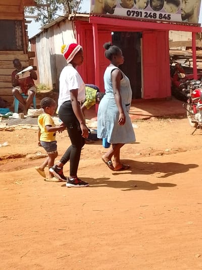 The scene is a dusty street in front of a barber shop, located in Lwandeti, Kenya (latitude: 0.563, longitude: 34.806). Four people are present. A young girl in a yellow shirt and blue shorts walks in the foreground, slightly to the left. She is being led by a woman with a red and white hat, wearing black pants, and a white t-shirt. The woman has dark, curly hair and is walking briskly. Behind them, a woman with short, dark hair is wearing a blue dress and walking in the opposite direction. This woman is glancing towards a man sitting on a bench in the background, wearing a blue shirt and tan shorts. The man is looking at a book or magazine. The shop’s exterior is a bright pink, with a sign that reads "0791 248 845". The shop sign also features photographs of men with various hairstyles. There is a motorcycle parked in the background.