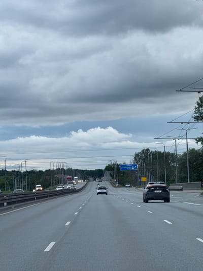 The image captures a view from inside a moving vehicle on a multi-lane highway under a cloudy, overcast sky. The highway is paved with asphalt and marked with white lane dividers. Several cars and trucks are visible on the road, all moving in the same direction. In the distance, there are signs indicating routes to "BAUSKA," "KAUNAS," and "VILNIUS." The highway is flanked by trees and numerous utility poles with overhead wires, suggesting an urban or suburban environment. The overall impression is of a typical commute on a day with poor weather conditions. The signage suggests the location is likely on the outskirts of Riga, Latvia, as these are major routes leading out of the city.