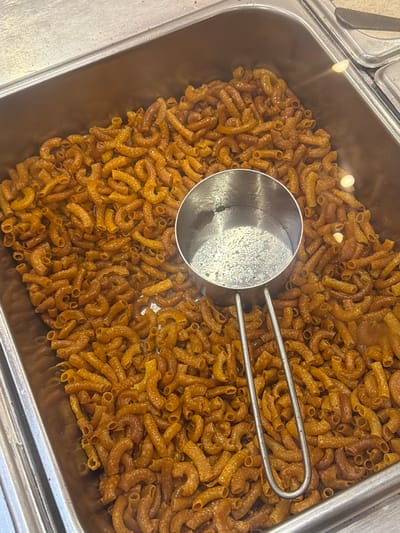The image shows a close-up, overhead view of a metal tray filled with macaroni. A metal scoop with a curved handle rests on top of the pasta. The macaroni pieces are curved and appear to be coated in a sauce or glaze, giving them a shiny, golden-brown appearance. The tray itself is part of a larger serving setup, with another section visible to the left. The lighting suggests an indoor setting, possibly a food service establishment. The scene is still and focused on the food. No people, text, time of day, or weather details are discernible. The location context is East Jakarta, Indonesia.