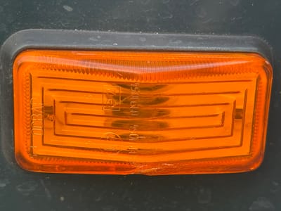 An orange plastic reflector is mounted on a dark grey surface. The reflector has a textured interior with horizontal lines and a raised border. Some text and numbers are visible embossed on the reflector, including "DCBAD", "E2", and "0101054". The dark grey surface appears to be painted metal, possibly part of a vehicle. The scene is an outdoor close-up, with some dust or dirt visible on both the reflector and the surrounding surface. The location is in the city of Sredny, Russia.