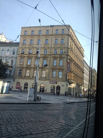 The image depicts a street scene in Prague, Czechia, likely from the perspective of a tram. The foreground shows tram tracks on a cobblestone street. Several buildings line the street, with the most prominent being a light yellow, multi-story building with ornate architectural details. This building features numerous windows and arched doorways at street level, housing businesses including KFC restaurants, identifiable by their logos and signage.
Several individuals are visible on the sidewalk in front of the buildings, appearing to be pedestrians walking or standing. The environment suggests a commercial or urban area, with streetlights and overhead wires for the tram system present. The sky is clear and blue, indicating daytime. The overall impression is a lively, urban setting in the historic city of Prague.