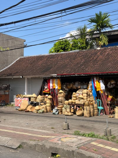 The image depicts a street-side market stall in Blahbatuh, Indonesia, filled with a variety of woven goods. The stall has a traditional tiled roof and is open to the street, displaying numerous woven baskets, hats, mats, and bags. In front of the stall, the items are arranged on the pavement and on low shelves. Some of the woven items are conical hats, while others are larger, round baskets. Behind the display of woven goods, there are colorful fabrics and umbrellas hanging, as well as more woven bags and other products stored within the stall. A bright blue plastic chair is visible to the right of the display. The street in front of the stall is paved with bricks and concrete, with a curb and a drainage grate. A sign with Indonesian text is propped up near the front of the stall. In the background, there are closed metal roller doors of a building, palm trees, and overhead electrical wires. The sky is clear and blue. There are no people visible in the image.