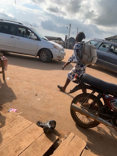 A young woman with braided hair wearing a blue and white floral dress and carrying a light-colored backpack walks along a dusty road. She is wearing dark, flat shoes. Beside her, a motorcycle is parked, its red body visible. The motorcycle has a sticker on its side that reads "Knock on the door...Open." Further down the road, a white minivan is stopped, with a driver visible inside. Behind the minivan, another dark-colored vehicle is partially visible. The scene appears to be a street in Shagamu, Nigeria, under a cloudy sky. The sunlight casts long shadows, suggesting it is either morning or late afternoon. The ground is covered in reddish-brown dirt, indicative of a dry season. Scattered on the ground are pieces of trash, including a black plastic bag. The overall atmosphere suggests a busy, perhaps traffic-laden, urban or semi-urban environment.