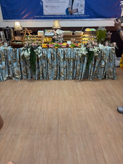 This image shows a buffet table set up indoors, likely for an event. The table is covered with a light blue floral-patterned tablecloth that is gathered at the bottom. On the table, there are various food items including what appears to be miniature sandwiches, a salad in a wooden bowl, a cake, fruit platters, chips, and a large dispenser filled with green liquid. Decorative flowers and greenery are arranged on and around the table. Behind the buffet table, a large blue banner with text is visible. The flooring is made of wood-look tiles. There are also two lamps with intricate shades on the table. The scene suggests a catered event or celebration. The presence of a banner from "The Medical City Pangasinan" indicates the location is likely in Pangasinan, and given the specific mention of Dagupan, Philippines, it is probable that this event is taking place in or near Dagupan City. No individuals are prominently featured in the image, though a hand holding a phone is partially visible on the right side.
