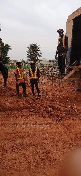 The image displays an outdoor scene, likely a construction site or undeveloped land, characterized by reddish-brown, dry, and cracked soil. The sky overhead is bright and appears overcast or lightly cloudy.
Several individuals, presumably workers, are present in the scene. On the far left, a person is standing near a dark, metallic structure, partially obscured, wearing a yellow or orange reflective vest. In the mid-ground, two men are standing upright. Both are wearing reflective vests over dark clothing and dark shoes; one has a yellow vest and the other a red/orange vest. The man in the yellow vest also appears to be wearing a hard hat or cap. Another individual is partially visible in the lower-left corner, dressed in dark clothing.
On the ground near the center, between the standing men and the large structure, is a small, dark, rectangular piece of equipment with wires extending from it. These wires run across the dry earth. In the background, beyond the immediate work area, there are glimpses of distant buildings and some green vegetation, including a prominent palm tree, suggesting the location is near a populated area.
The individuals appear to be engaged in some form of outdoor work or observation, given their attire and the presence of equipment on the ground. The overall impression is that of a daytime work scene in a dry, possibly tropical or arid environment. No discernible text is visible in the image.