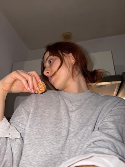 A fair-skinned woman with auburn hair in a messy bun holds a peeled tangerine in her hand, displaying manicured pink nails with glitter. She wears a grey sweatshirt layered over a white collared shirt. The background shows a simple interior with a white wall, a light fixture, and what appears to be a refrigerator or cabinet. The image is angled, presenting a distorted perspective.