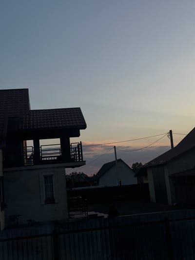 This image captures a somewhat eerie scene at dusk in Chemerne, Ukraine. The photo, taken vertically, shows the silhouette of several buildings against a pale sky streaked with the muted colors of sunset. Power lines stretch across the frame, adding to the urban, yet somewhat desolate, atmosphere.
The main subjects are the buildings themselves. On the left, a dark, tiled roof juts out, a stark contrast to the smooth, gray sky. Next to it, a darker, more complex structure rises, featuring open-air levels with railings and window openings. Further to the right, another building with a white facade and a rectangular window is visible. The right side of the image is dominated by the dark, textured surface of a wooden or metal wall, possibly part of a larger building or fence.
The time of day is clearly late evening or early dusk, with the sky transitioning from daylight to night. The weather appears calm, with no visible signs of rain or wind. The location is indicated as Chemerne, Ukraine. There are no people or specific events depicted; the focus is on the architecture and the ambient light. The overall mood is quiet, perhaps even melancholic, due to the dim lighting and the stark architectural forms. No text is visible in the image.