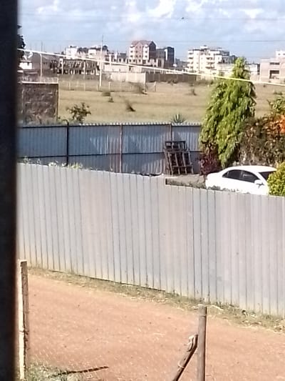 The image depicts a daytime scene in Kalimoni ward, Kenya, characterized by a mix of undeveloped land and urban development under a partly cloudy sky.

In the foreground, a reddish-brown dirt path or unpaved ground is visible, bordered by a chain-link fence with two visible wooden posts on the left and right. Immediately behind this, a tall, light gray corrugated metal fence extends horizontally across the frame.

In the mid-ground, a second, slightly lower corrugated metal fence parallels the first. Behind this, to the left, is a visible section of a stone-walled structure or unfinished building. The central area features a large expanse of unkempt, dry grassy land with scattered small trees and shrubs. To the right, behind the lower corrugated fence, are several stacked wooden pallets. Adjacent to the pallets, a dense green conifer-like tree partially obscures the rear portion of a white sedan parked behind the fence line.

The background reveals an elevated landscape transitioning into an urban environment. Multiple multi-story buildings, some appearing to be under construction or newly built, form a skyline against the light blue sky with scattered white clouds. The overall scene suggests ongoing development and expansion. No individuals are visible.