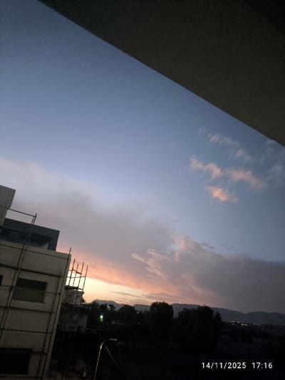 The image, captured on 14/11/2025 at 17:16, presents a twilight scene over Nicosia, Northern Cyprus / Cyprus. The sky transitions from a deep blue at the top to a prominent orange and pink at the western horizon, indicative of a sunset, with scattered clouds reflecting these colors. A dark, angled architectural element, likely a balcony ceiling or building overhang, occupies the upper third of the frame. In the left mid-ground, a multi-story building under construction is visible, featuring a light-colored facade and extensive scaffolding. The central and right mid-ground consists of dark, silhouetted trees and vegetation. A distinct green light source and several fainter, distant lights are visible within this vegetated area. The background features a silhouette of distant mountains or hills spanning the horizon, adorned with multiple clusters of small lights, indicating an urbanized or populated area. No individuals or specific human actions are depicted.