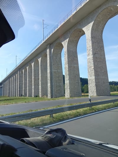 The image shows a massive stone viaduct with numerous arches, stretching across a landscape under a clear blue sky. The viaduct appears to be a railway bridge, as indicated by the overhead catenary lines visible above its deck. In the foreground, a portion of a car's interior is visible, including a dark-colored dashboard and a cap resting on it. The perspective suggests the photo was taken from inside a vehicle traveling alongside the viaduct, which is a prominent feature of the Rizenbach, Switzerland landscape. The viaduct's stone construction is detailed, with individual blocks clearly visible, and its scale is emphasized by the repetitive pattern of arches receding into the distance. The surrounding environment includes a grassy verge, a road, and some trees and fields in the background.