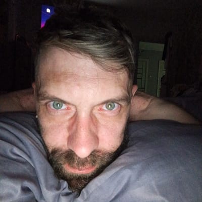 A close-up, dimly lit selfie of a man with a beard and light green eyes. The man is lying down, possibly on a bed, with his arms outstretched behind his head on either side. A gray pillow or bedding is visible beneath his face. Redness is apparent in the corners of both eyes. A television screen is visible in the background. A slightly ajar, white door with a hanging dark garment is in the background. The location is Марковский, Russia.
