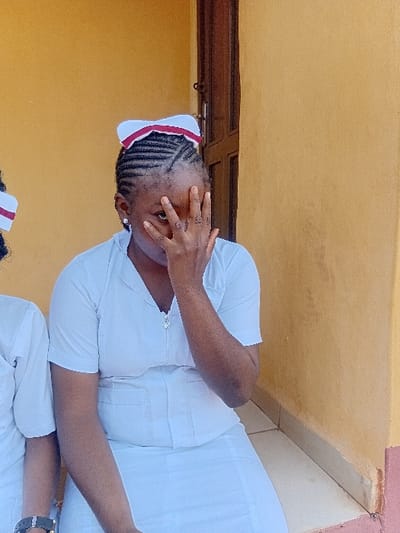 An adult female of African descent is depicted in Ejule, Nigeria. She is wearing a white short-sleeved uniform with a V-neck and a metallic zipper pull, and a matching white skirt. A white cap with a red band is worn on her head, over dark cornrowed hair. Her right hand covers the lower half of her face, partially obscuring her nose and mouth, with her left eye looking directly at the camera. A small earring is visible in her left ear. To her immediate left, the partial figure of another individual, also in a white uniform, is visible, including a left wrist adorned with a dark-strapped watch. The background consists of a plain, warm yellow-orange wall. A dark brown wooden door frame is partially visible vertically on the right side of the frame. The subjects appear to be seated, with a tiled floor visible in the lower foreground, featuring light-colored tiles transitioning to a reddish-brown base.