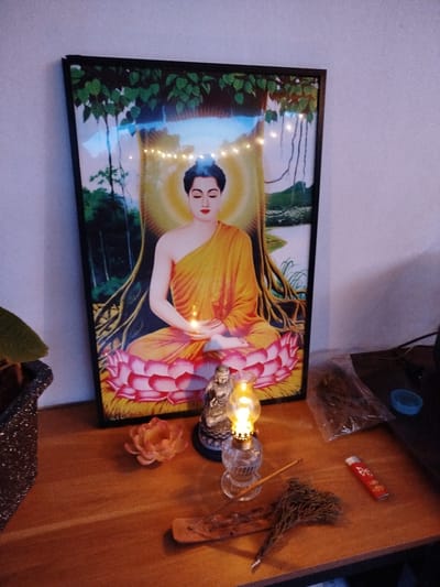 The image depicts a devotional altar set up in what appears to be a home environment in Lạc Dương, Vietnam. The central subject is a framed picture of the Buddha meditating under a Bodhi tree, with a glowing aura and a lotus flower base. String lights are draped over the top of the frame, adding a decorative touch.
In front of the picture, a small Buddha statue sits beside a miniature oil lamp, its flame casting a warm glow. A single unlit incense stick rests on a wooden stand, with a bundle of dried herbs or incense material nearby. A small, decorative lotus flower sits to the left of the incense. To the right, a red lighter and a clear plastic bag filled with dried plant material are visible. A small blue cap or container is also present. A plant in a textured pot is partially visible on the far left.
The lighting suggests it might be evening or a dimly lit indoor space, creating a serene and contemplative atmosphere. The overall scene evokes a sense of spirituality and personal devotion. No visible text is present in the image.